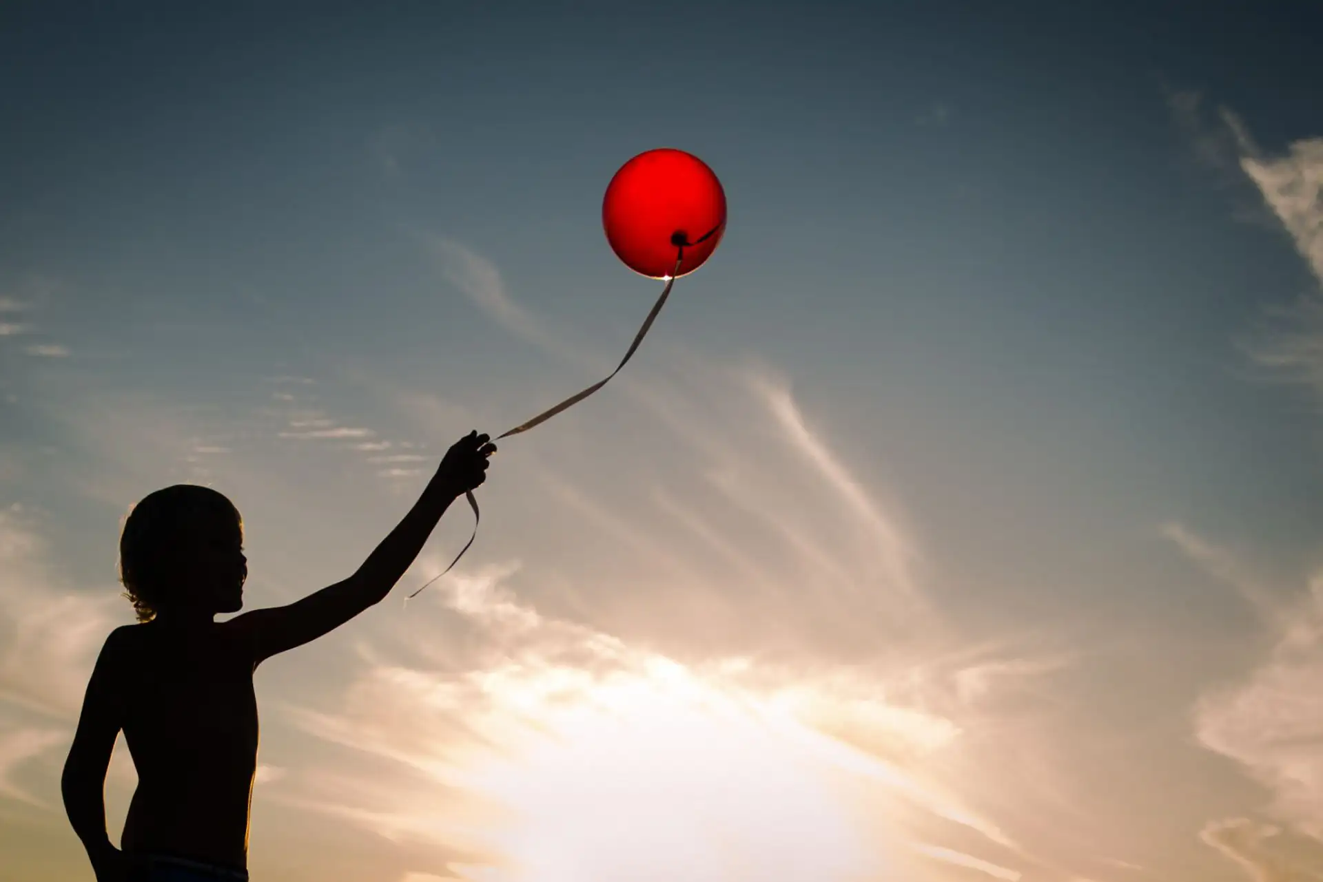 Kind hält roten Ballon gegen den Himmel bei Sonnenuntergang. Symbol für Freiheit.