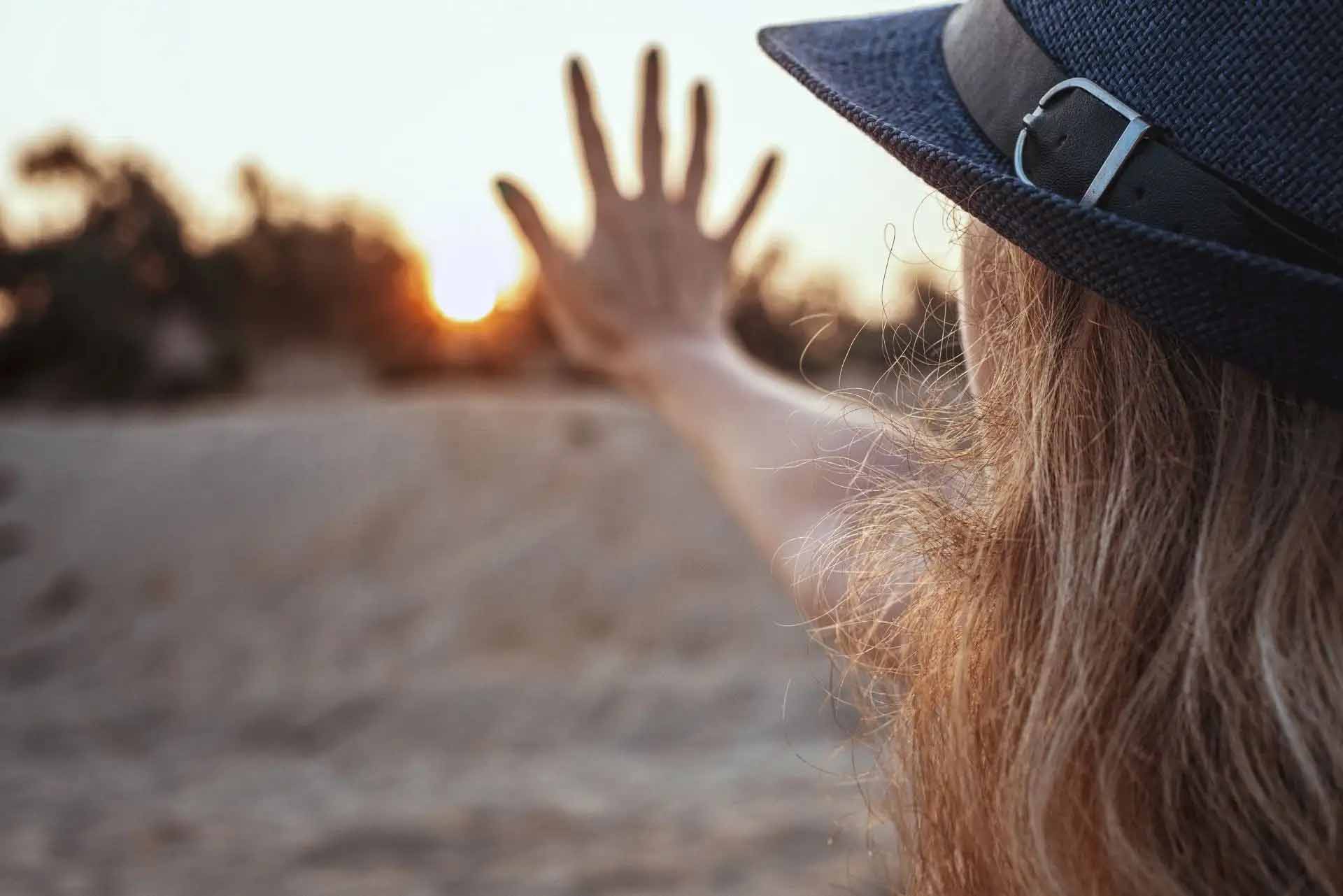 Frau mit Hut streckt Hand zum Sonnenuntergang, Natur im Hintergrund.