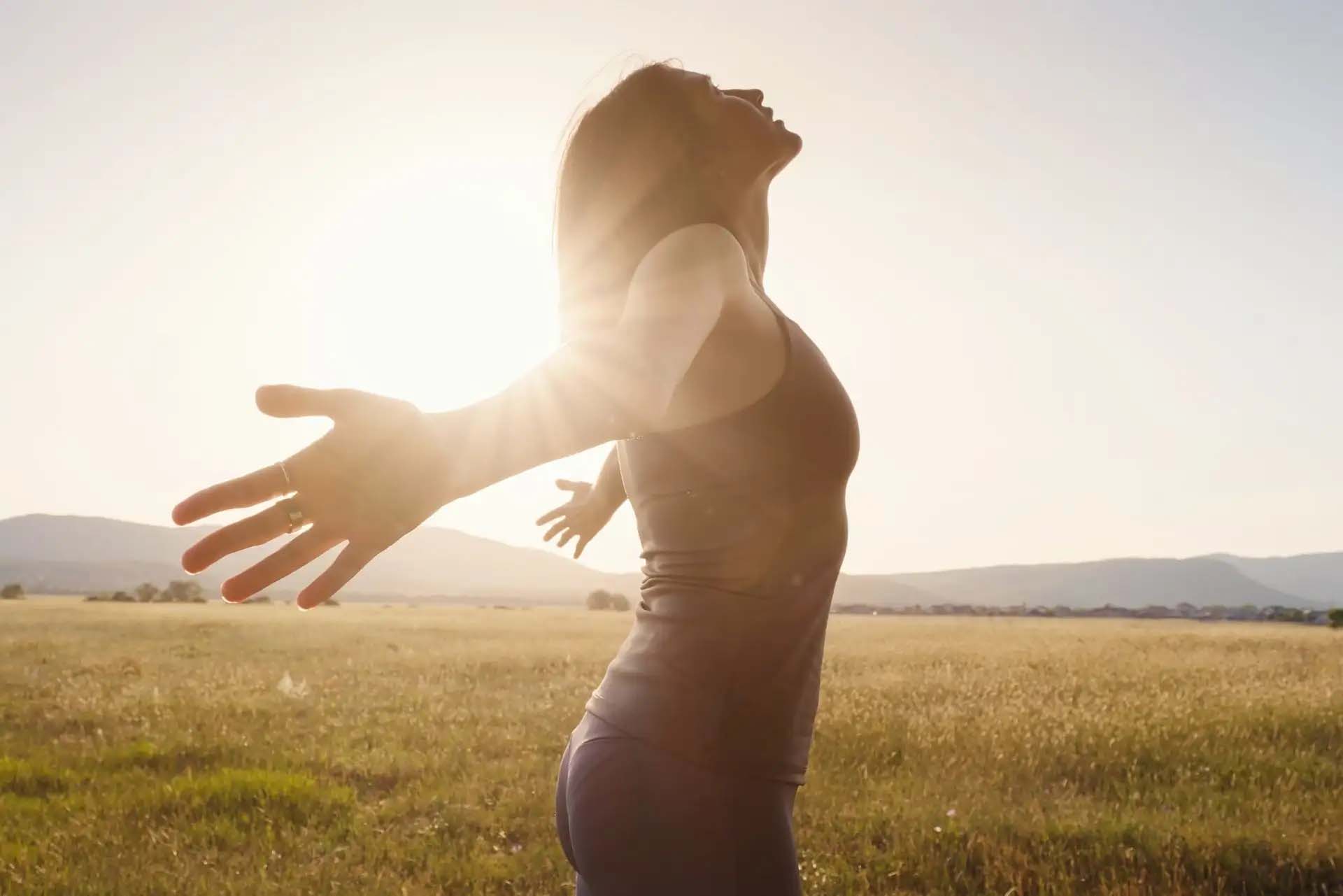 Frau steht mit ausgebreiteten Armen in der Sonne, fühlt Freiheit und Ruhe.
