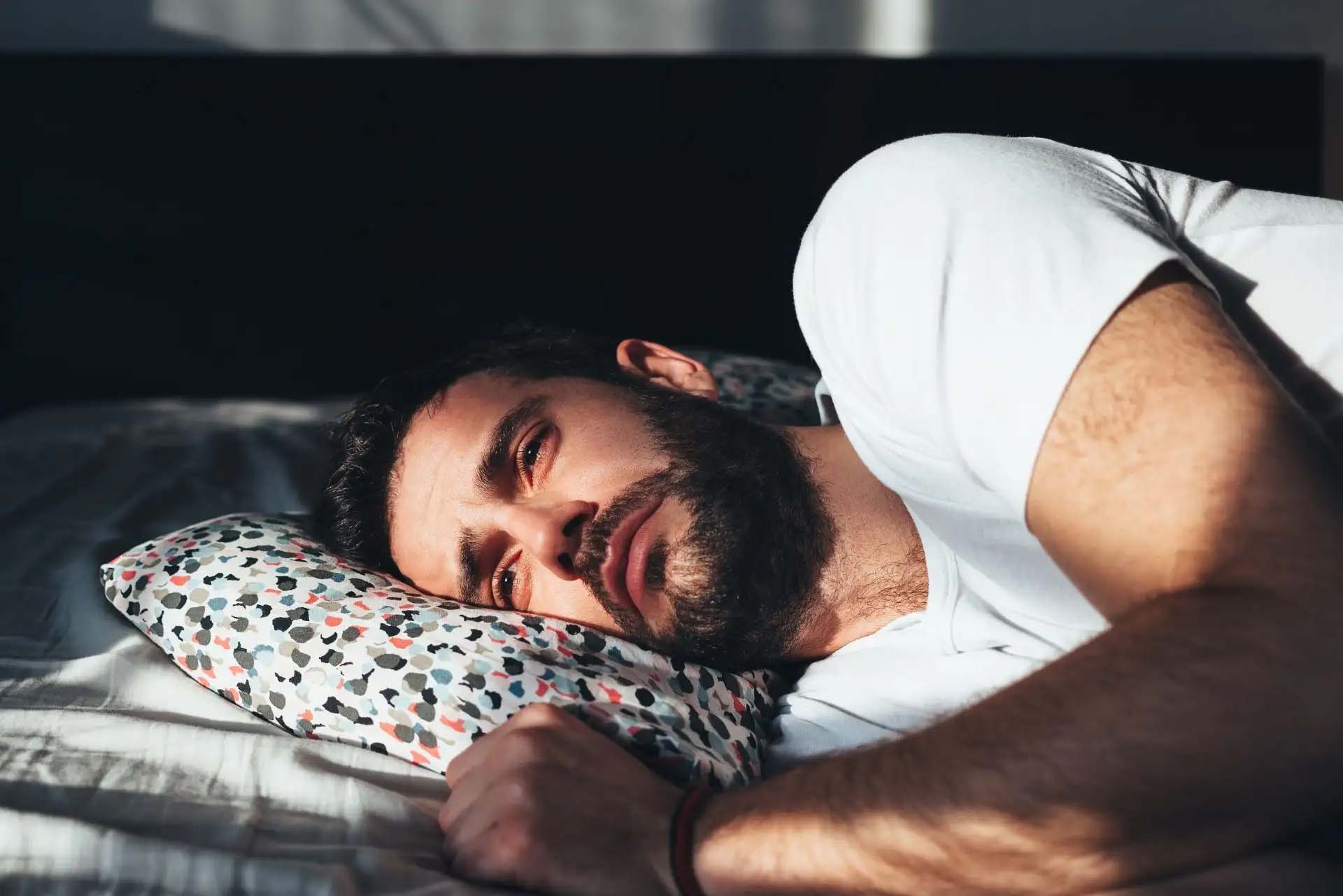 Ein nachdenklicher Mann liegt auf einem Bett, von Sonnenlicht beleuchtet.