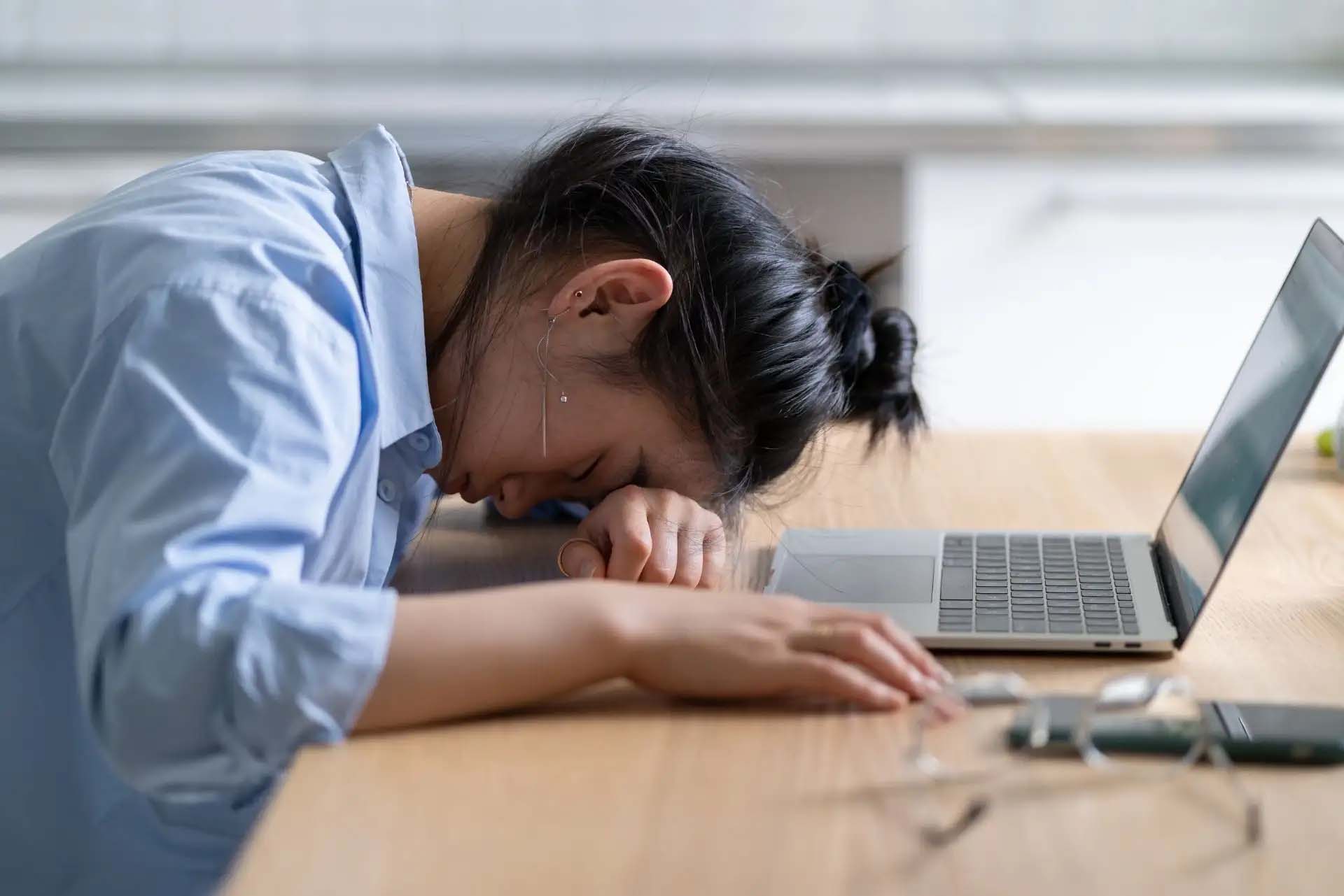Eine erschöpfte Frau legt ihren Kopf auf den Tisch neben einem Laptop.