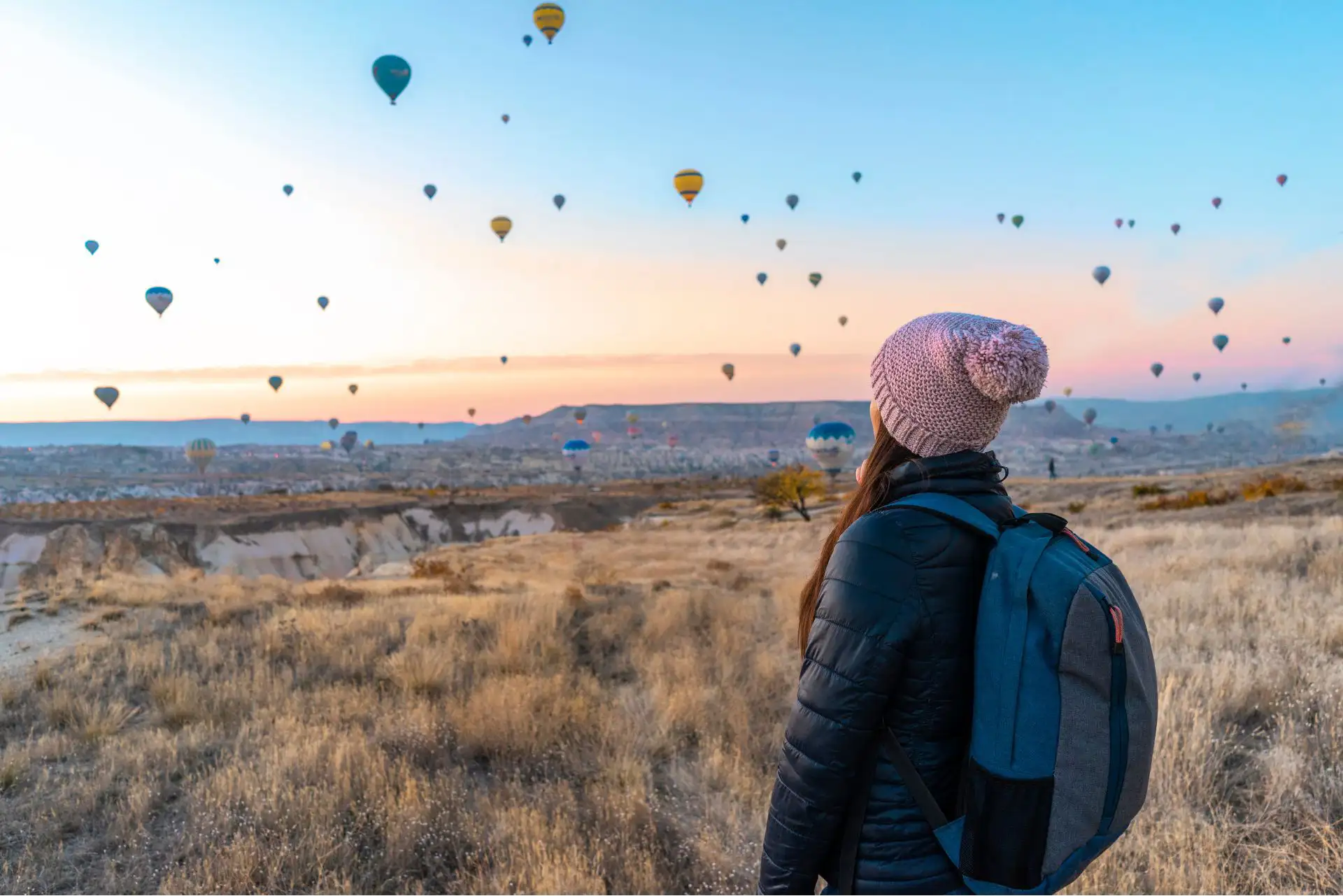 Eine Frau betrachtet zahlreiche Heißluftballons bei Sonnenaufgang über einer Landschaft.