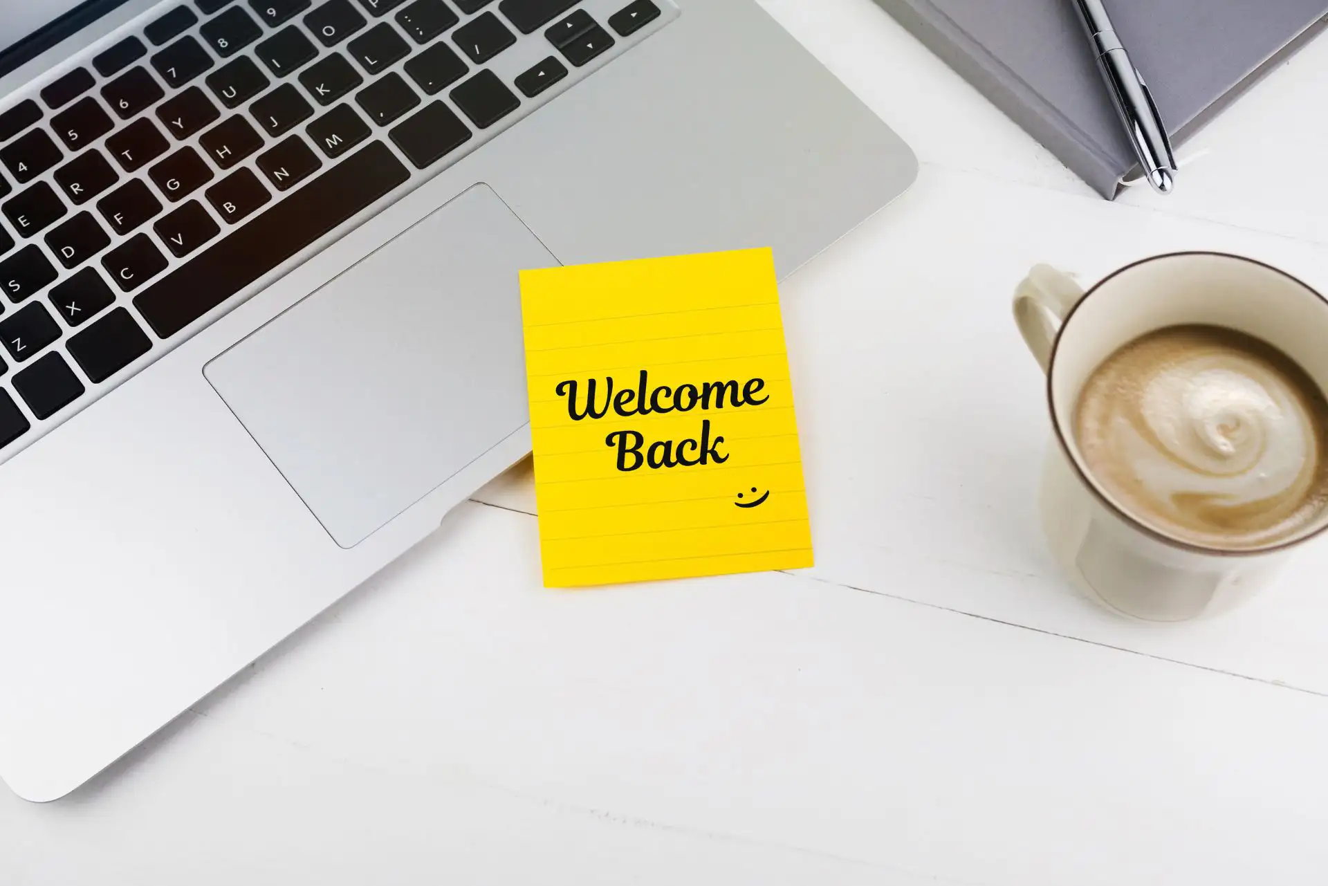 Gelber Notizzettel mit "Welcome Back" neben Laptop und Kaffeetasse.