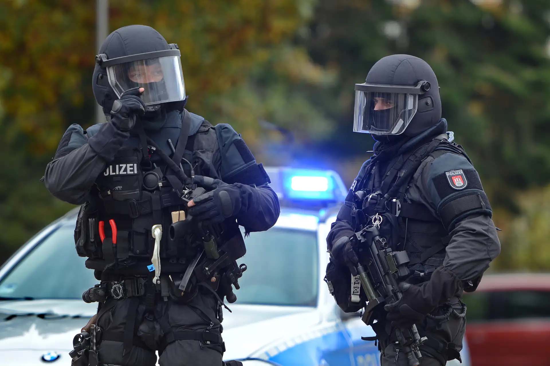 Zwei schwer bewaffnete Polizisten in Einsatzkleidung vor einem Polizeiwagen mit Blaulicht.