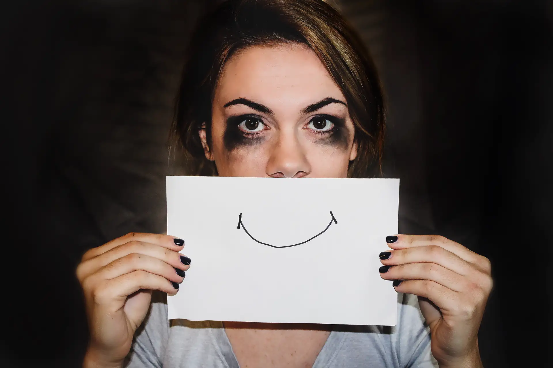 Frau mit verschmiertem Make-up hält Papier mit aufgemaltem Lächeln vor Gesicht.