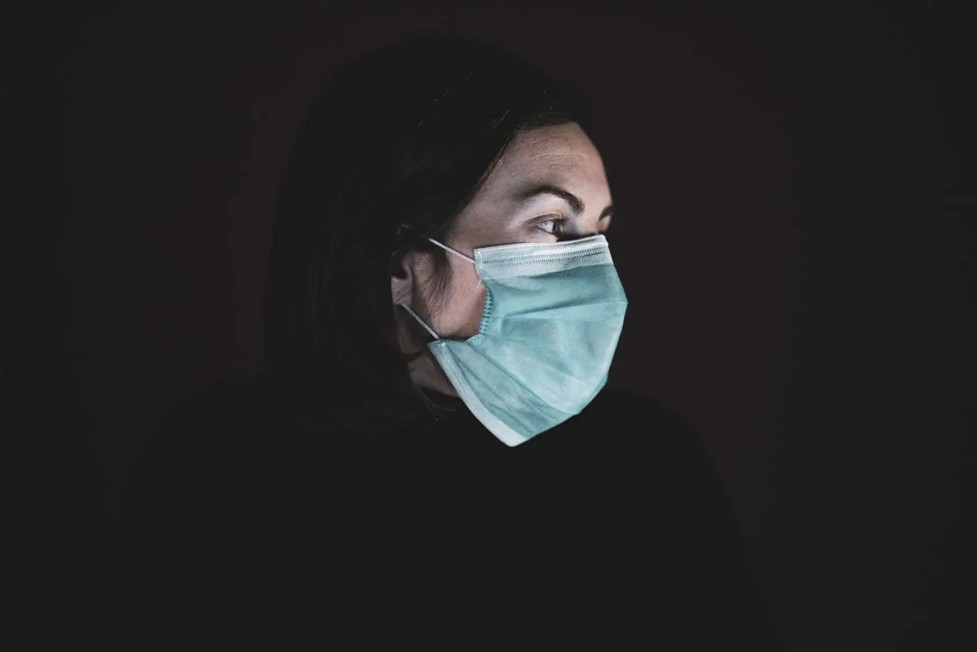 Eine Frau mit Maske blickt nachdenklich zur Seite, düstere Atmosphäre.