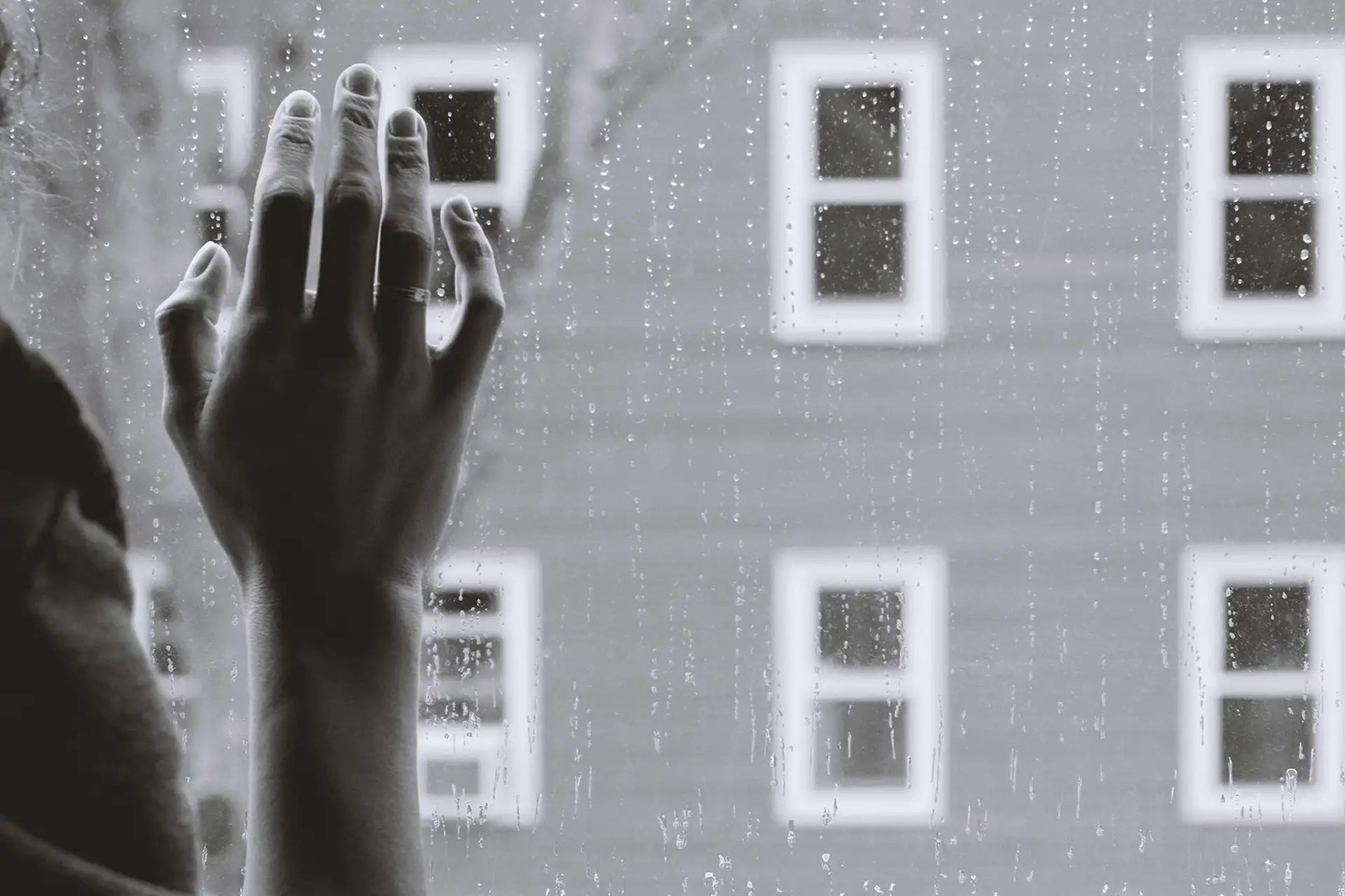 Hand einer Person liegt an regennassem Fenster, draußen graues Gebäude sichtbar.