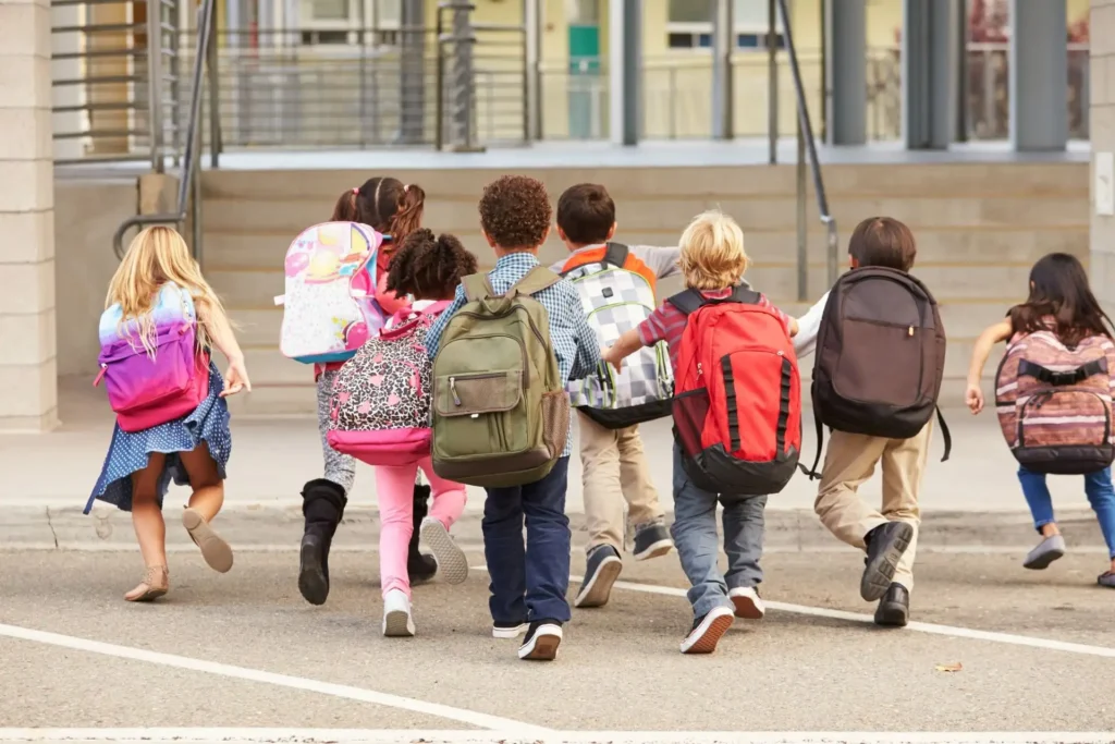 Kinder laufen mit bunten Rucksäcken fröhlich in Richtung Schuleingang.