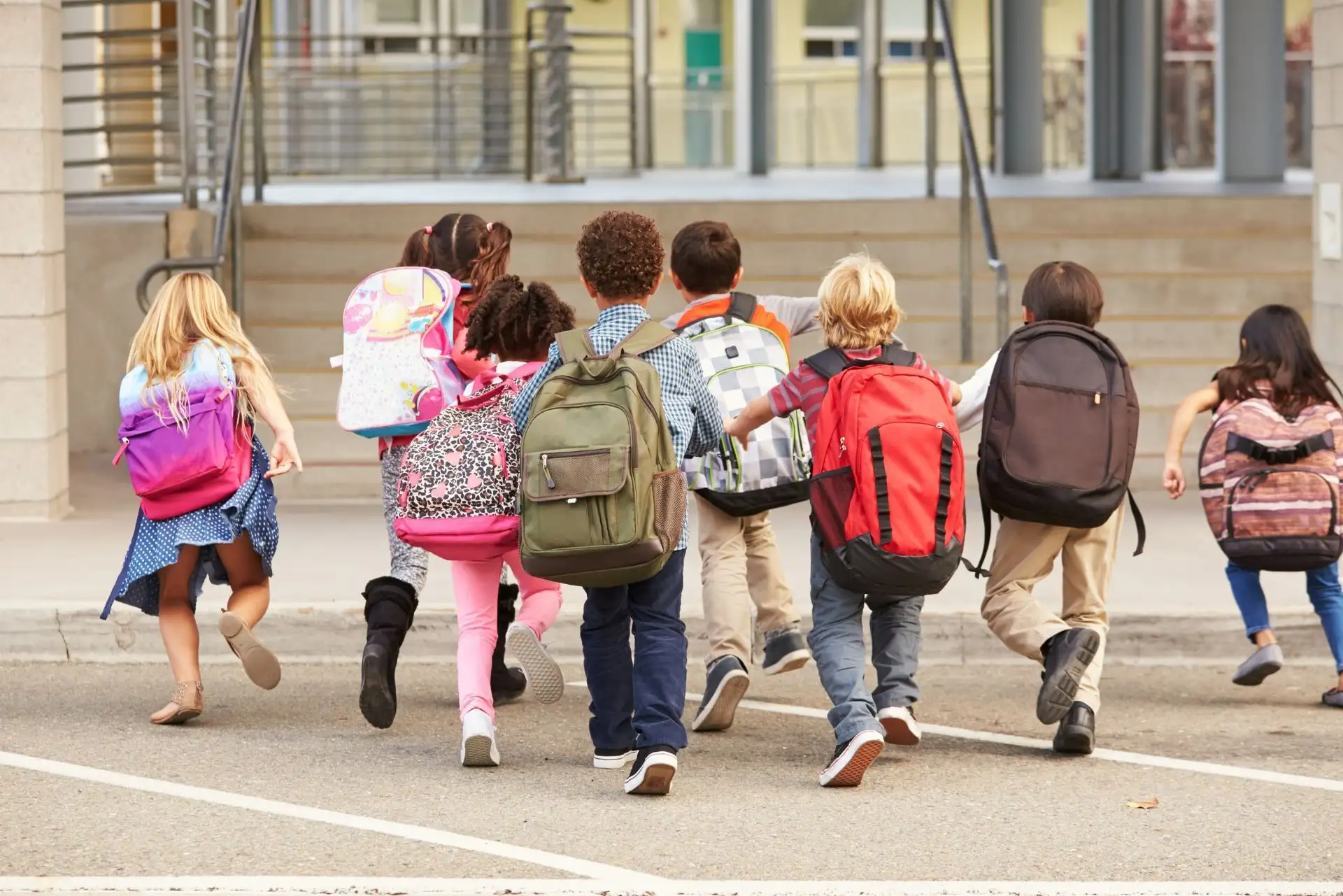 Kinder laufen mit bunten Rucksäcken fröhlich in Richtung Schuleingang.