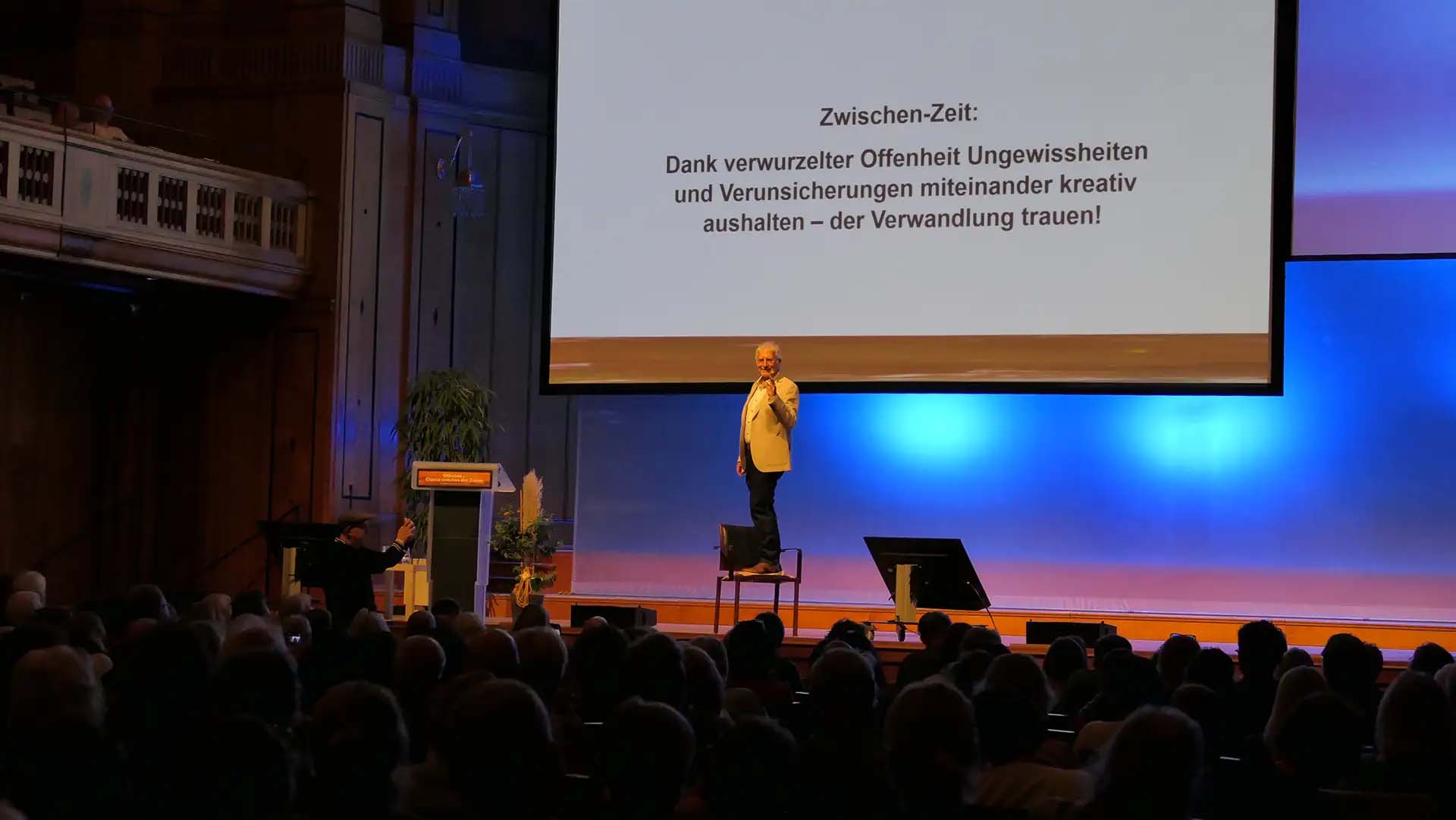 Theologe Pierre Stutz steht am Heiligenfeld Kongress 2024 auf der Bühne auf einem Stuhl.