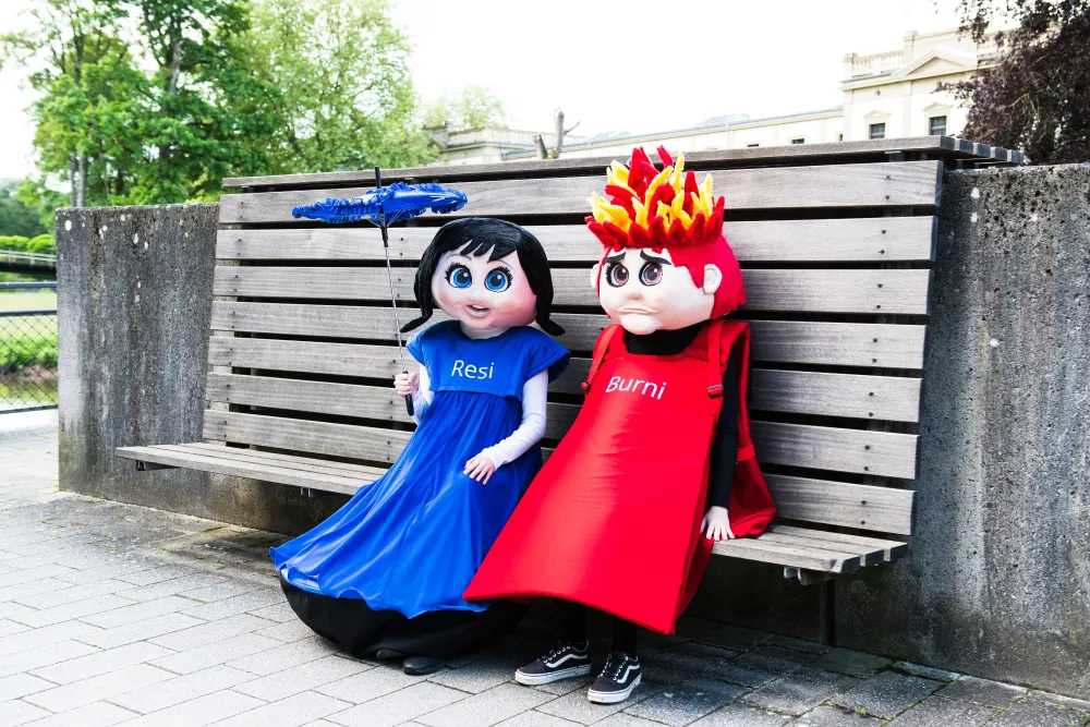 Maskottchen Resi und Burni sitzen auf einer Bank, Resi hält einen blauen Schirm.
