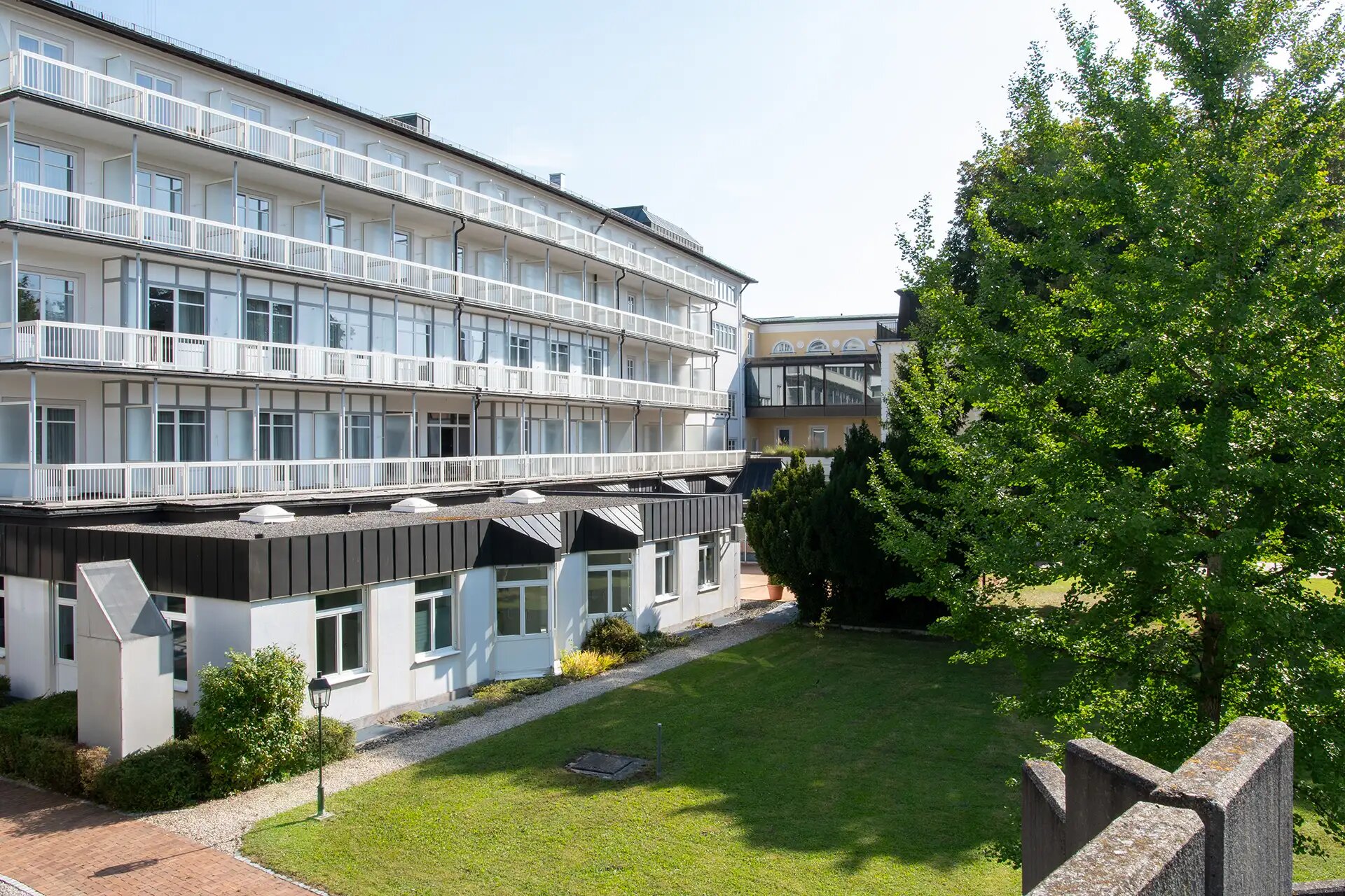 Modernes Gebaeude mit Balkonen und gruener Gartenanlage bei sonnigem Wetter.