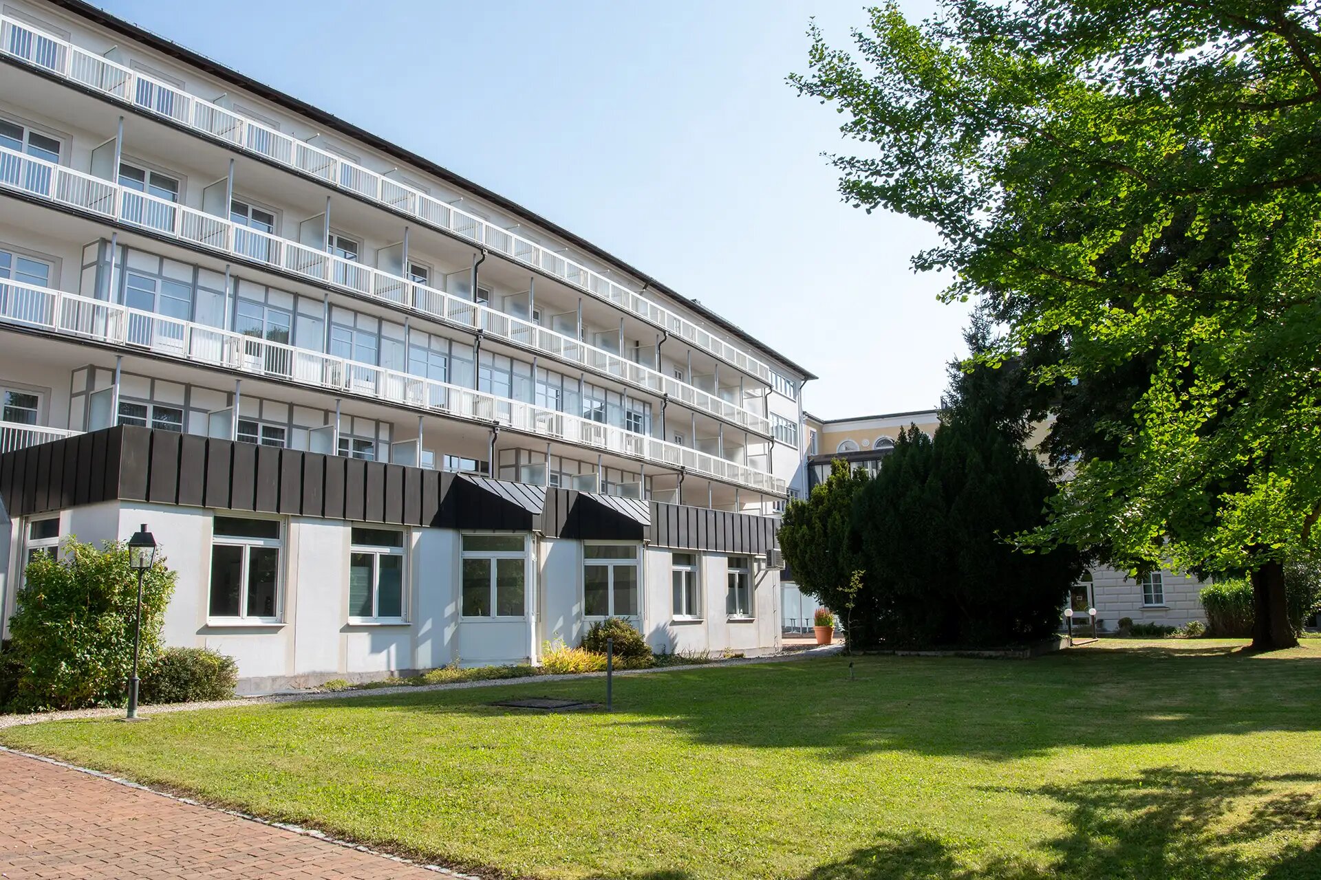 Modernes Gebaeude mit Balkonen und gruener Gartenanlage bei sonnigem Wetter.