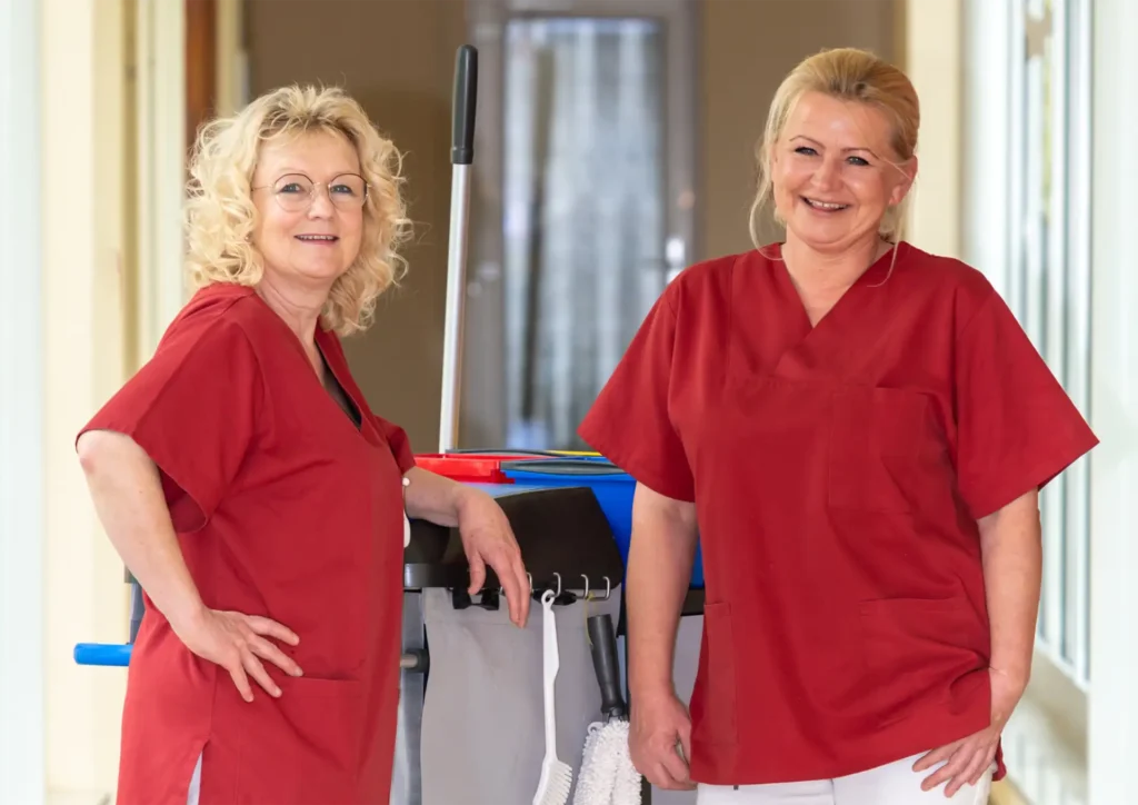 Zwei Reinigungskräfte in roter Arbeitskleidung stehen mit Reinigungswagen vor großen Fenstern.