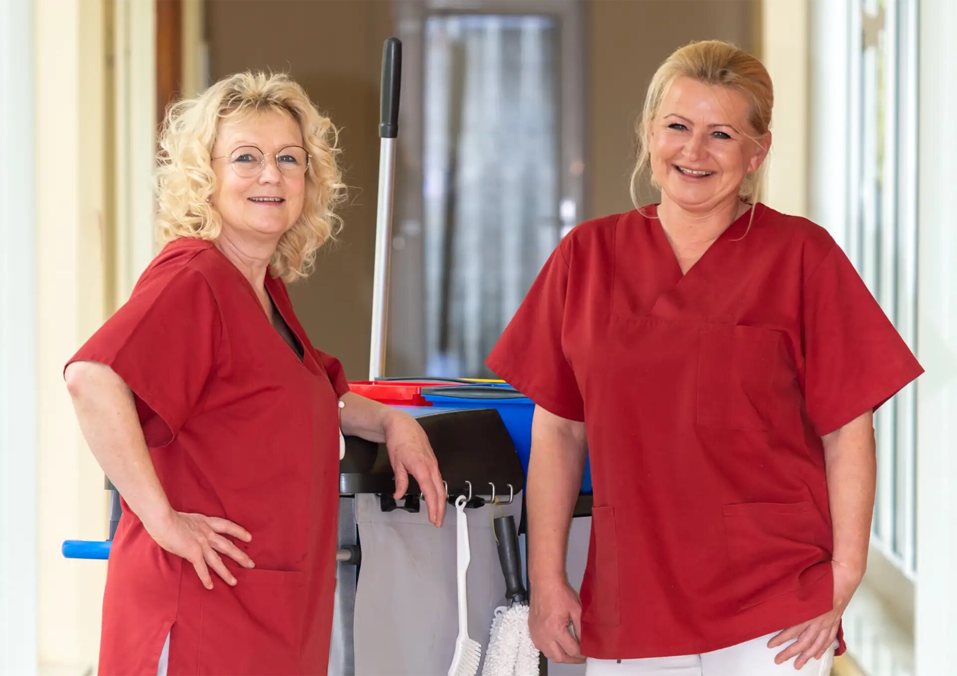 Zwei Reinigungskräfte in roter Arbeitskleidung stehen mit Reinigungswagen vor großen Fenstern.