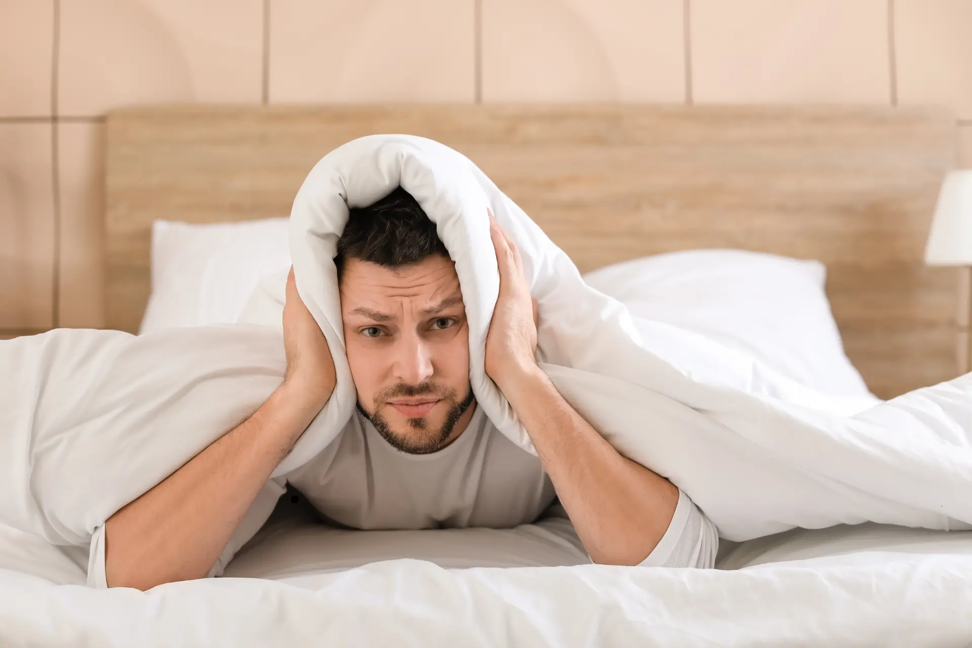 Ein Mann liegt auf einem Bett und hält sich mit der Bettdecke die Ohren zu.