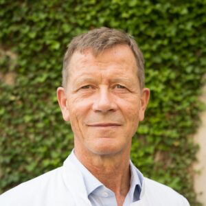Ein Mann mit weißen Arztkittel vor grünem Hintergrund lächelt freundlich