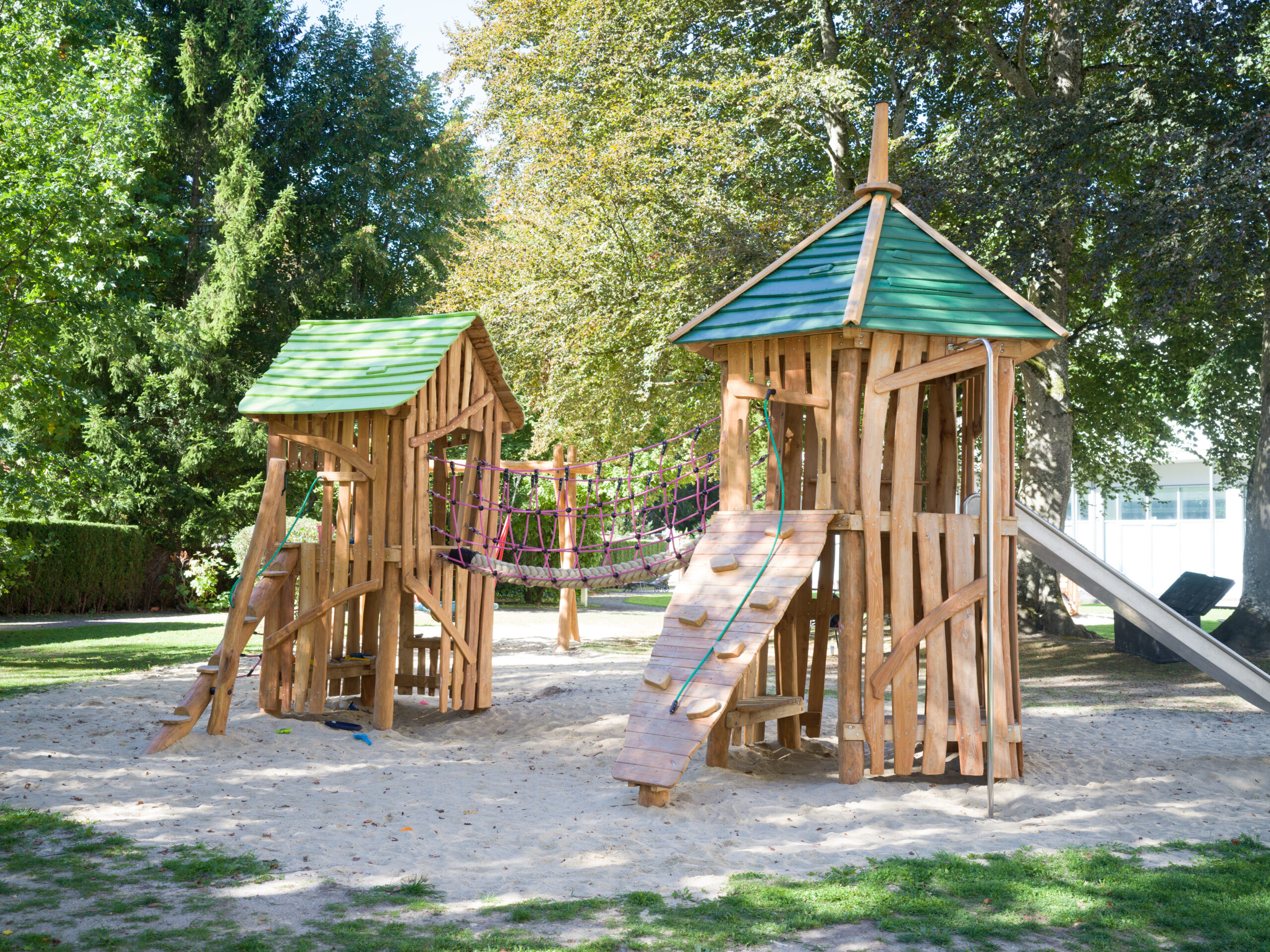 Spielgerüst für Kinder mit Kletterwand, Rutsche und Hängebrücke vor grüner Umgebung